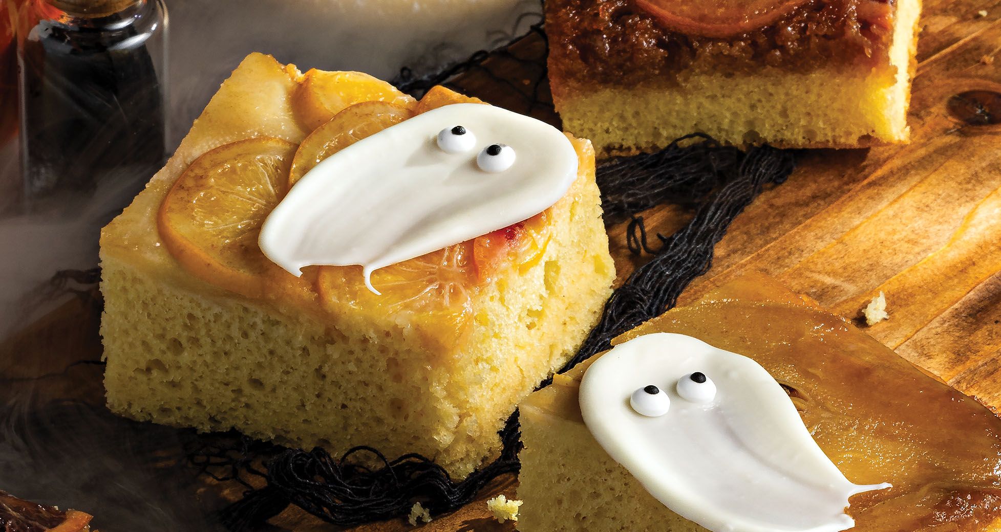 Square slices of citrus upside-down cake decorated with white icing ghosts with candy eyes for Halloween, served on a wooden board.