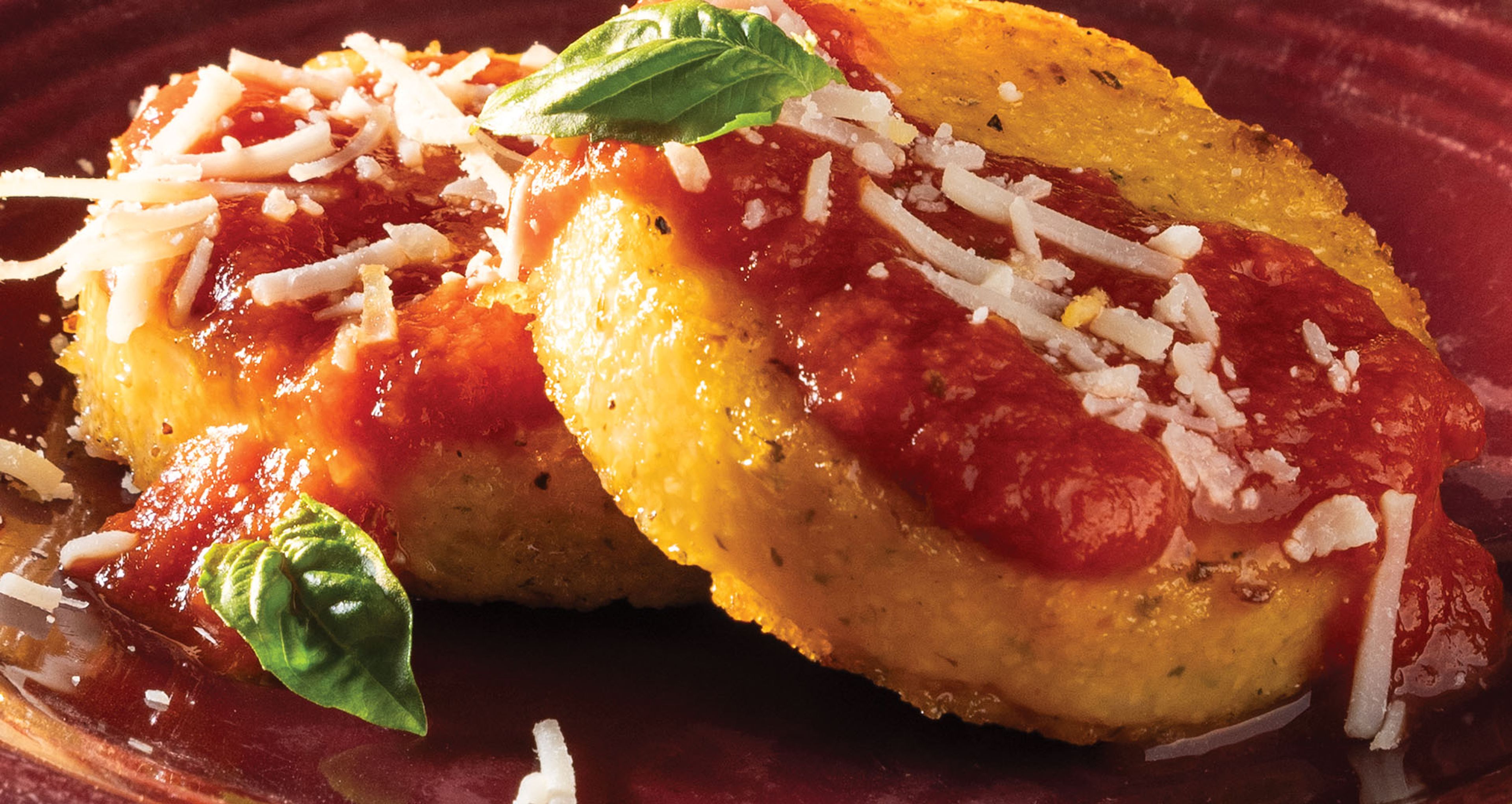 Two round pan-fried polenta cakes topped with red tomato sauce, shredded cheese, and fresh basil on a dark red plate.