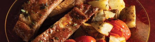 Glazed pork ribs served with roasted potato wedges and cherry tomatoes on a clear red glass plate.