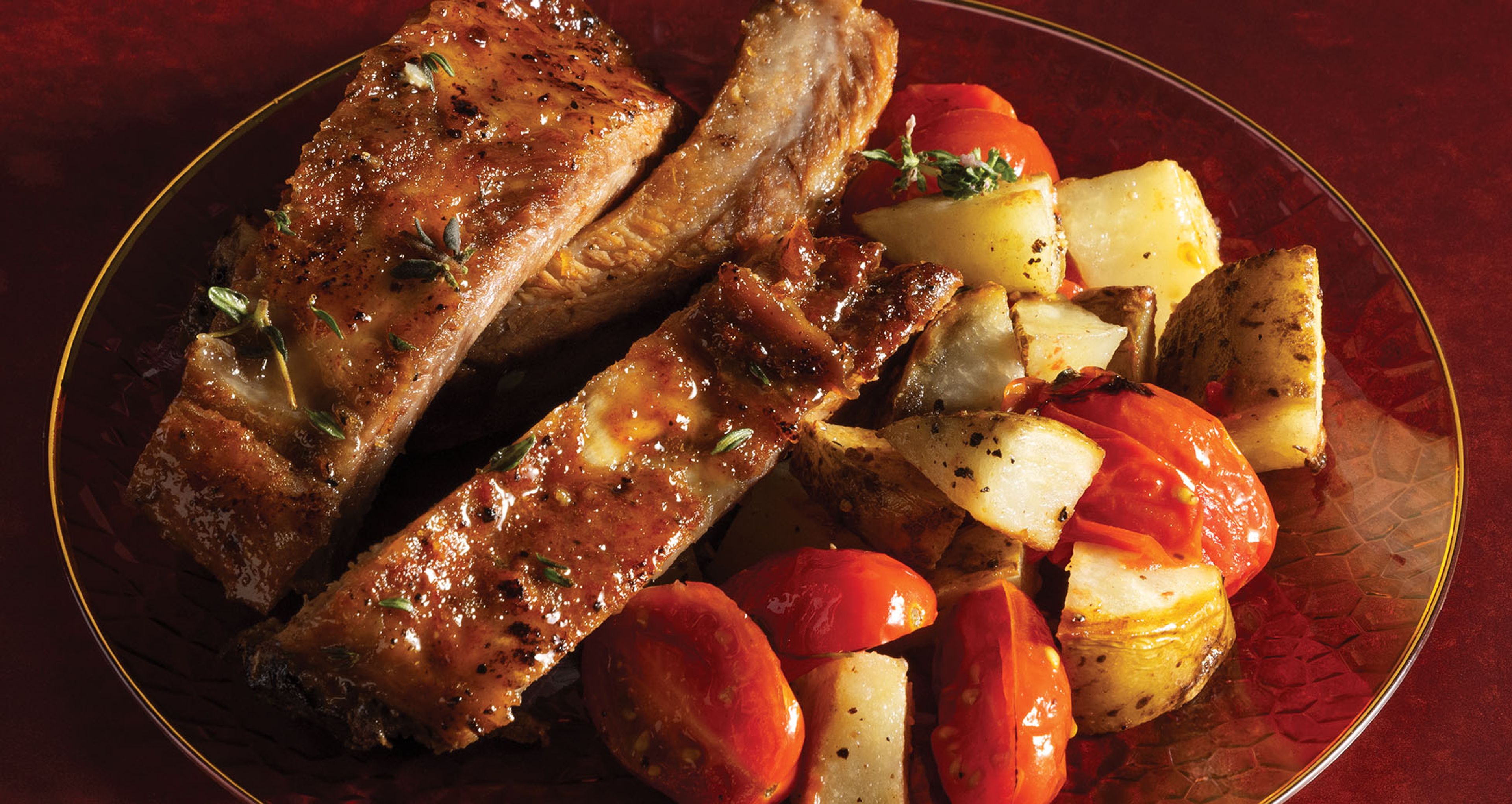 Glazed pork ribs served with roasted potato wedges and cherry tomatoes on a clear red glass plate.