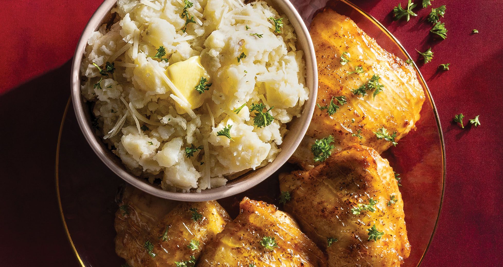 A dinner plate featuring glazed roasted chicken thighs served alongside a bowl of mashed potatoes garnished with butter, shredded cheese, and chopped parsley.
