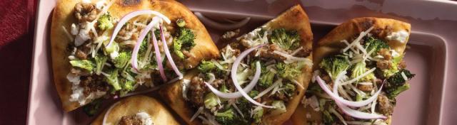 Four flatbread slices arranged on a light pink serving platter, topped with sausage, roasted broccoli, goat cheese, shredded cheese, and sliced red onion.