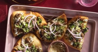 Four flatbread slices arranged on a light pink serving platter, topped with sausage, roasted broccoli, goat cheese, shredded cheese, and sliced red onion.