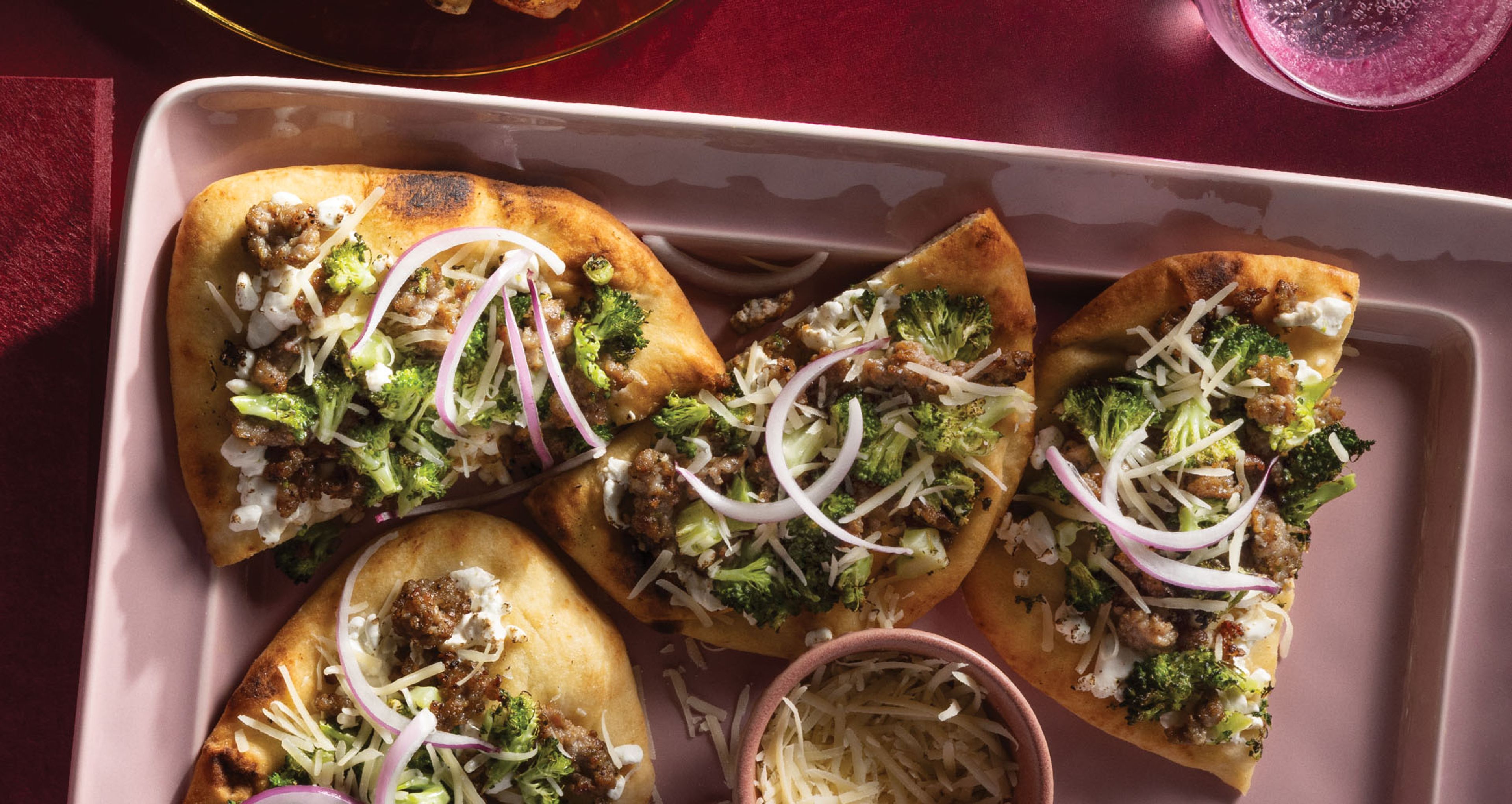Four flatbread slices arranged on a light pink serving platter, topped with sausage, roasted broccoli, goat cheese, shredded cheese, and sliced red onion.