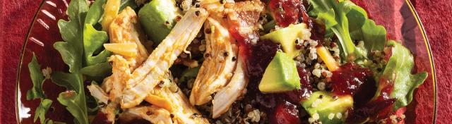 A chicken and quinoa salad with arugula, diced avocado, shredded cheddar, bacon, and cranberry sauce served on a red glass plate.