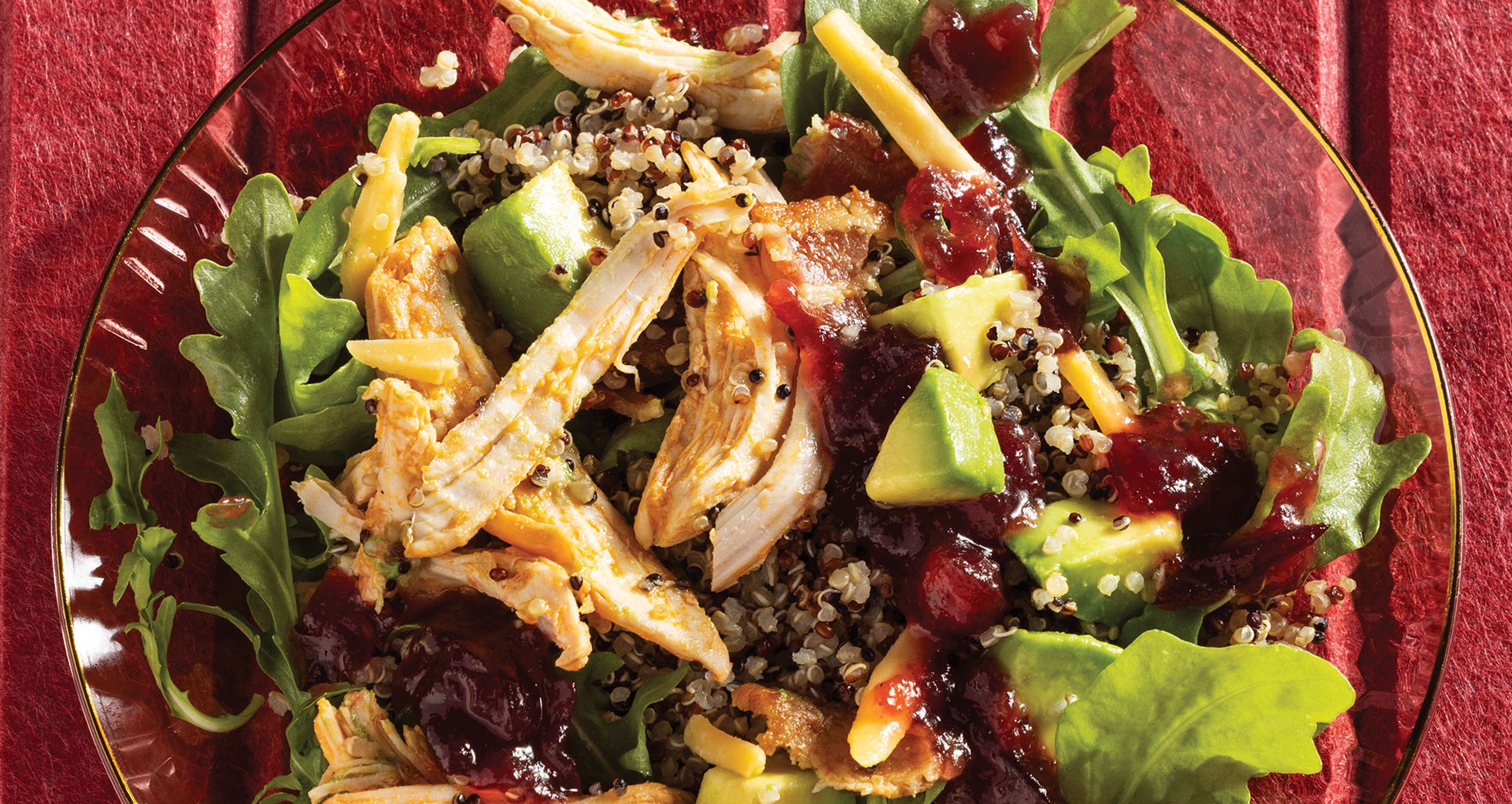 A chicken and quinoa salad with arugula, diced avocado, shredded cheddar, bacon, and cranberry sauce served on a red glass plate.