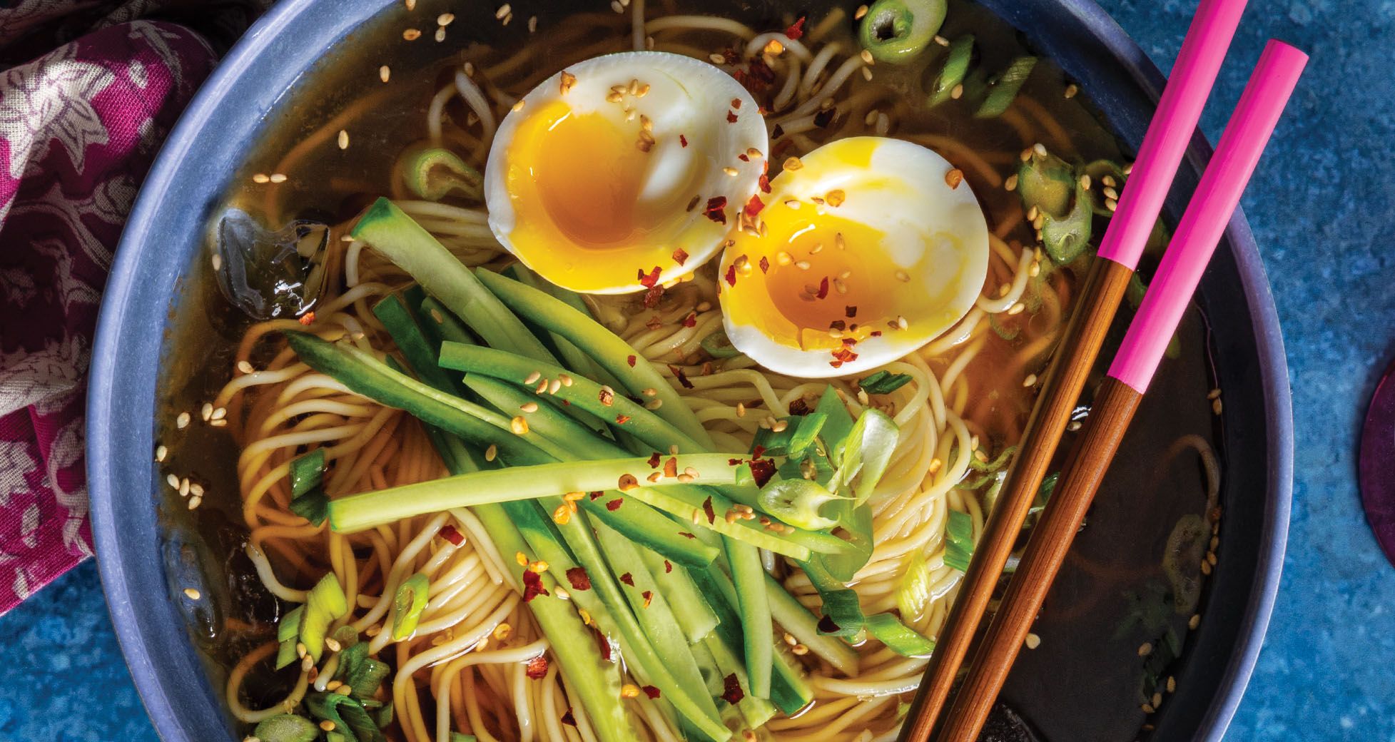 A bowl of ramen with soft-boiled eggs, sliced cucumber, scallions, chili flakes, and sesame seeds, paired with wooden chopsticks with bright pink ends.