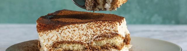A slice of tiramisu on a white plate with cocoa powder dusted on top and a fork lifting a bite above it.