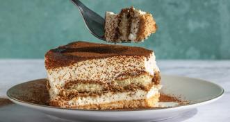 A slice of tiramisu on a white plate with cocoa powder dusted on top and a fork lifting a bite above it.