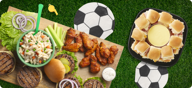 A wooden board with hamburgers, pasta salad, and chicken wings and a plate of sandwich sliders on a green turf background with napkins that look like soccer balls.