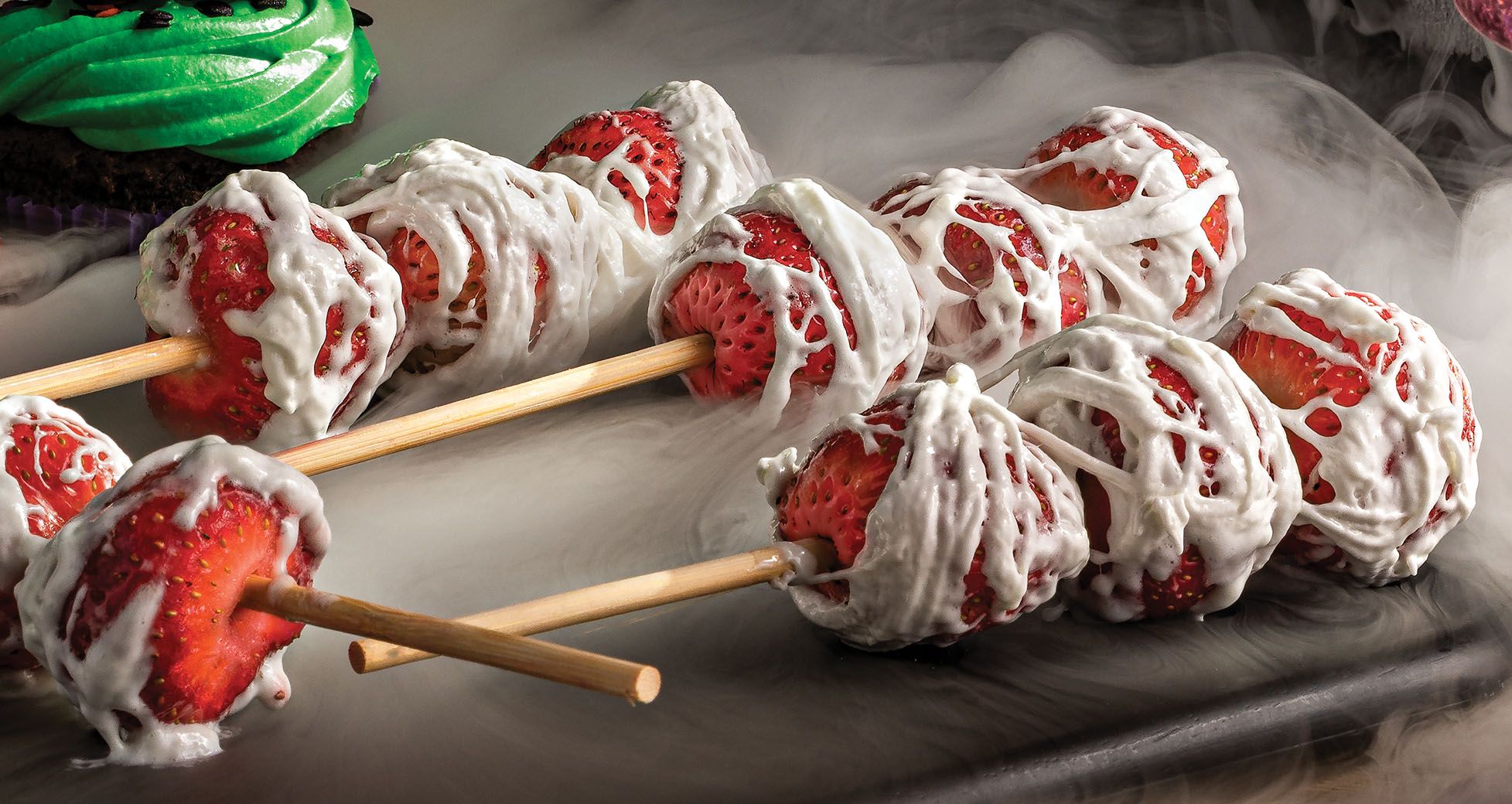 Strawberry skewers wrapped in marshmallow web icing for a Halloween theme, surrounded by white fog.