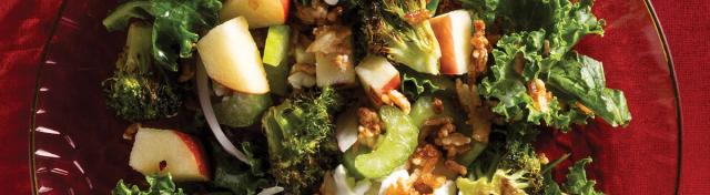 A colorful kale salad served on a red glass plate with apple chunks, roasted broccoli florets, celery, goat cheese, red onion, and crispy rice grains.
