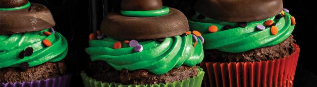 Three Halloween witch hat cupcakes topped with green frosting, festive sprinkles, chocolate cookie rims, and Hershey's kisses against a dark background.