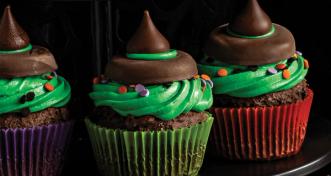 Three Halloween witch hat cupcakes topped with green frosting, festive sprinkles, chocolate cookie rims, and Hershey's kisses against a dark background.