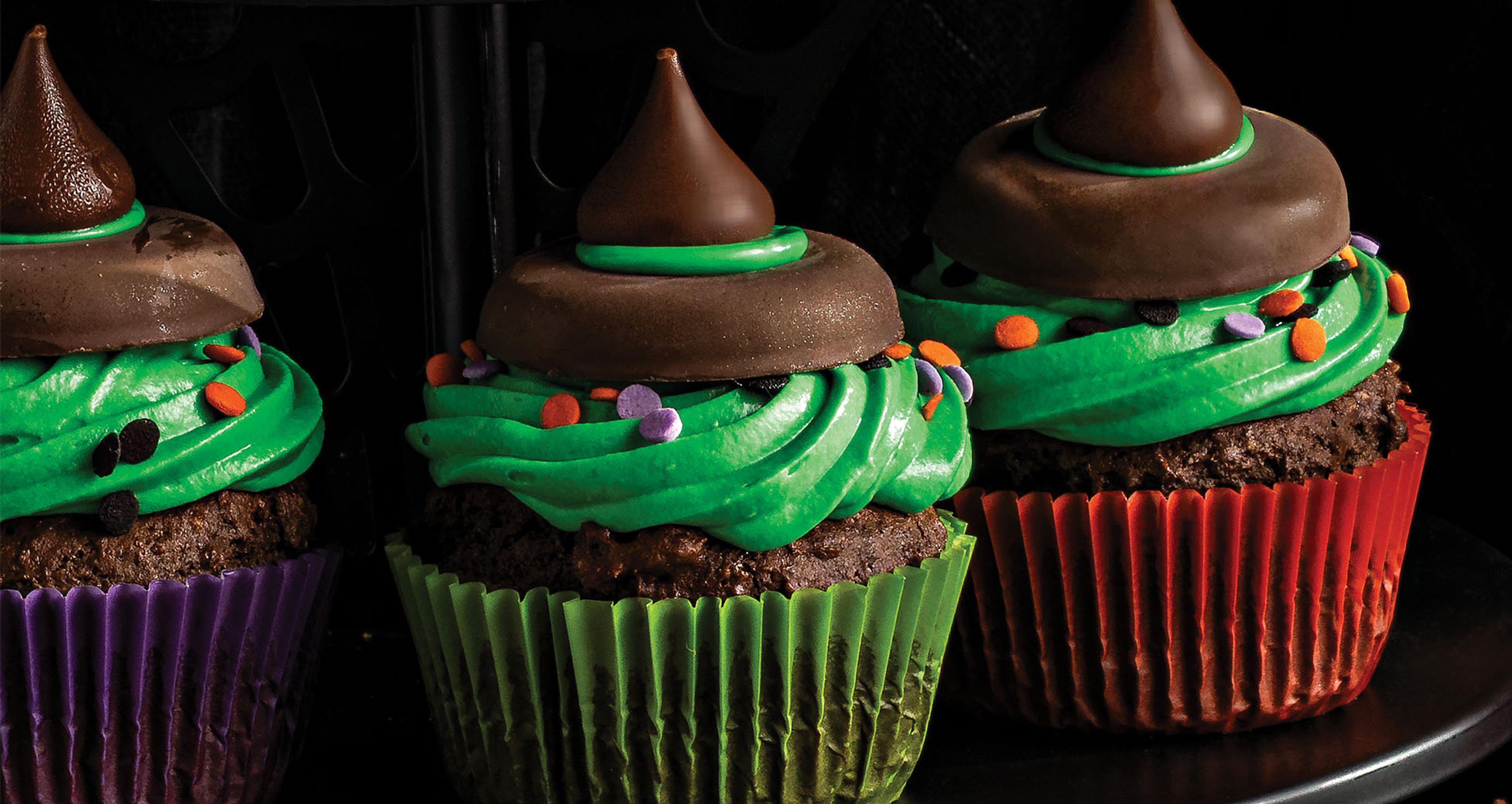 Three Halloween witch hat cupcakes topped with green frosting, festive sprinkles, chocolate cookie rims, and Hershey's kisses against a dark background.