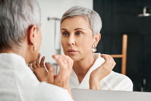 senior-woman-with-moisturizer-on-her-face-looking-at-mirror-in-picture-id1308206096