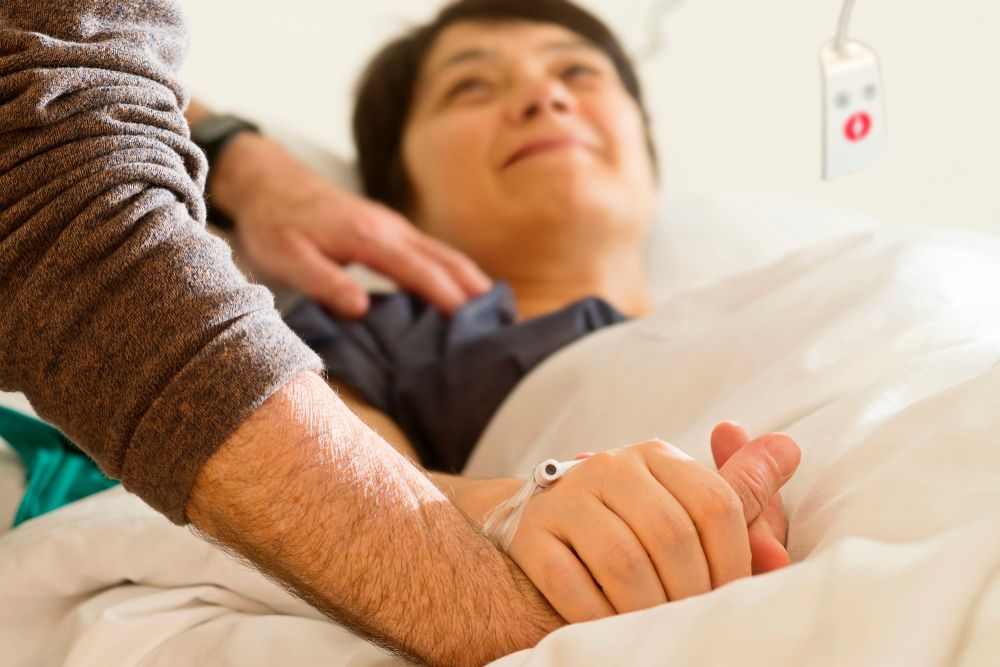 A woman on the hospital bed is holding her partner's hand
