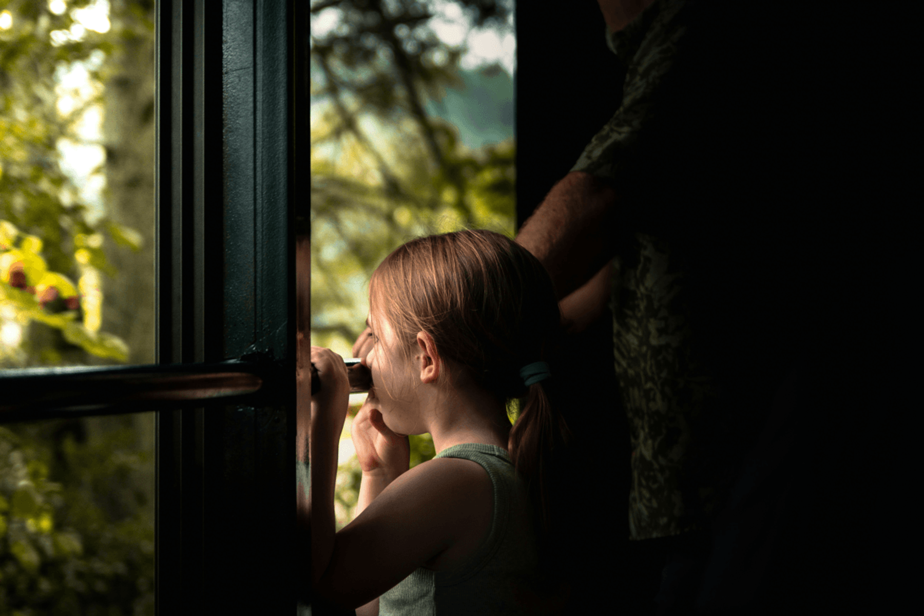 A young girl peering through binoculars out of a dark doorway into a sunlit forest, with an adult standing nearby.
