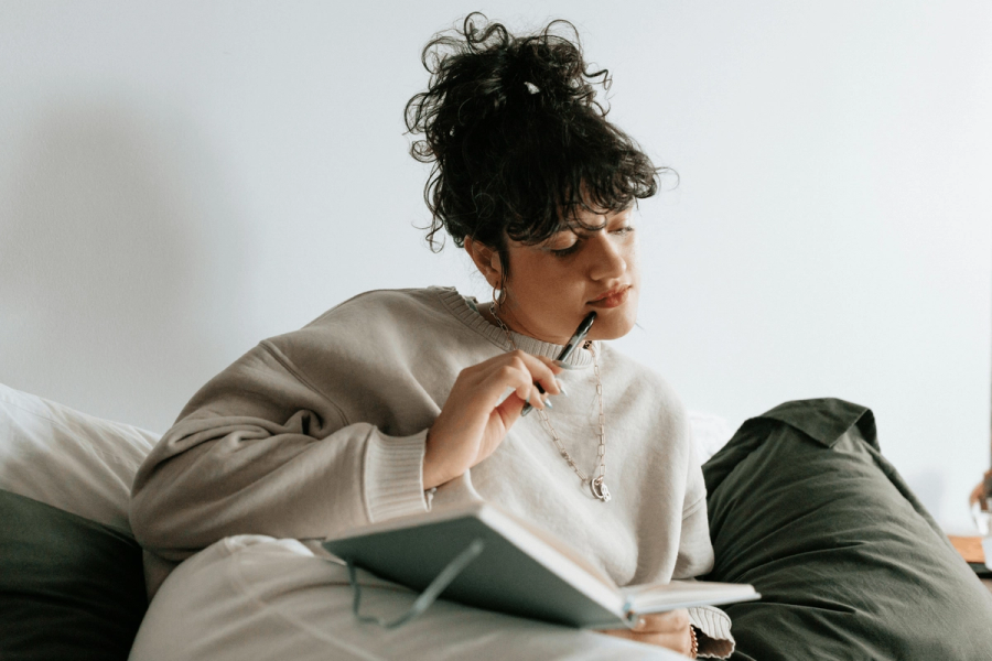 A young woman with curly hair in an updo lying on a bed, pensively holding a pen to her lips while writing in a journal.