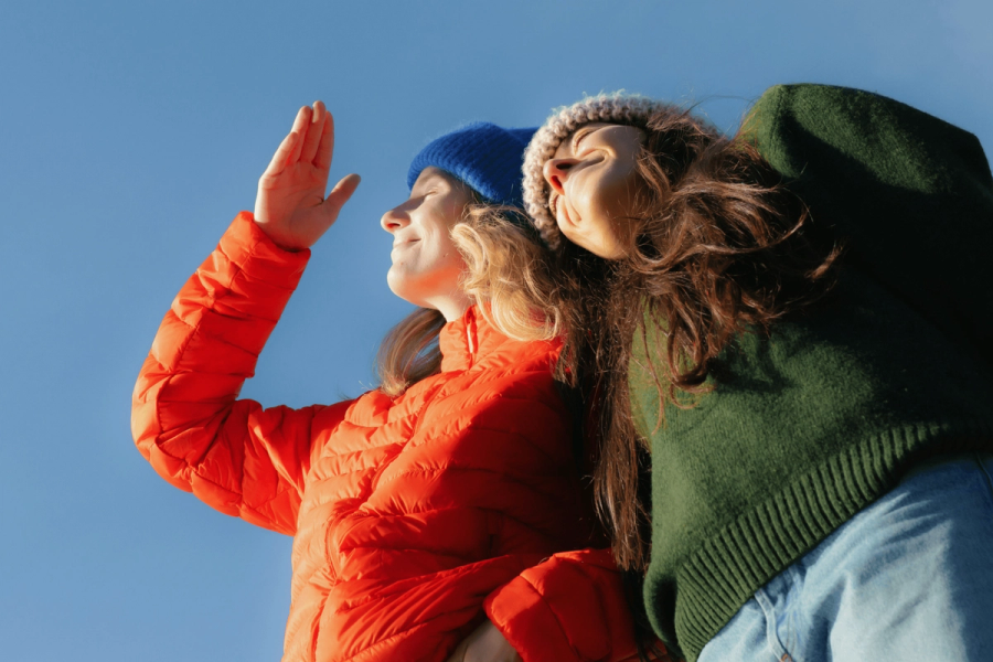 Two young women standing outdoors on a clear sunny day, faces tilted upward toward the sky with eyes closed in a peaceful, worshipful posture. One wears a bright orange puffer jacket and blue beanie, hand raised upward; the other wears a green sweater and knit hat, long hair blowing in the wind. Shot from a low angle against a vivid blue sky.