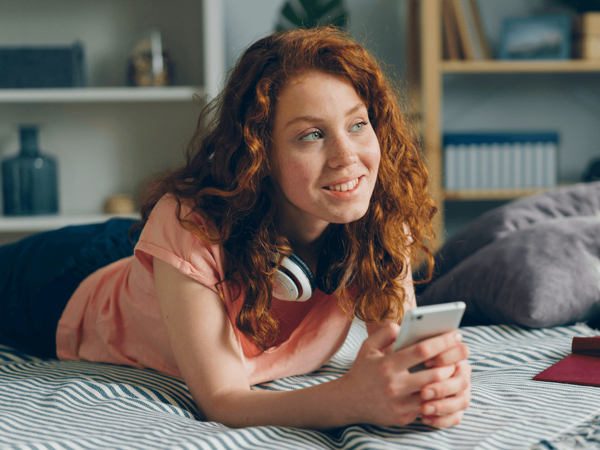 A young woman with curly red hair lying on a bed, holding a phone and smiling while looking to the side, with headphones around her neck.