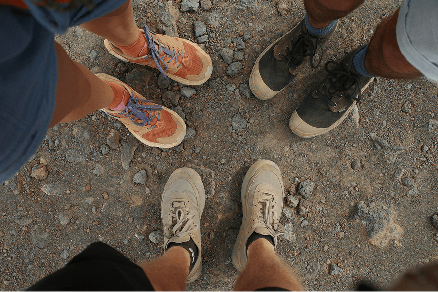A top-down view of three people's feet standing together on rocky ground, each wearing different trail shoes or hiking boots.