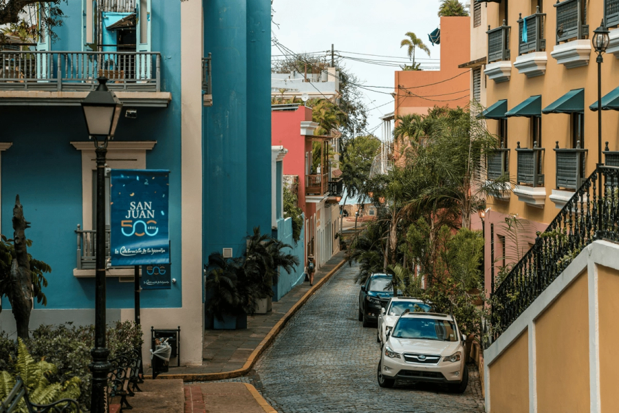 A colorful cobblestone street in Old San Juan, Puerto Rico, lined with vibrant colonial buildings in blue, pink, and yellow, with a San Juan 500th anniversary banner visible.