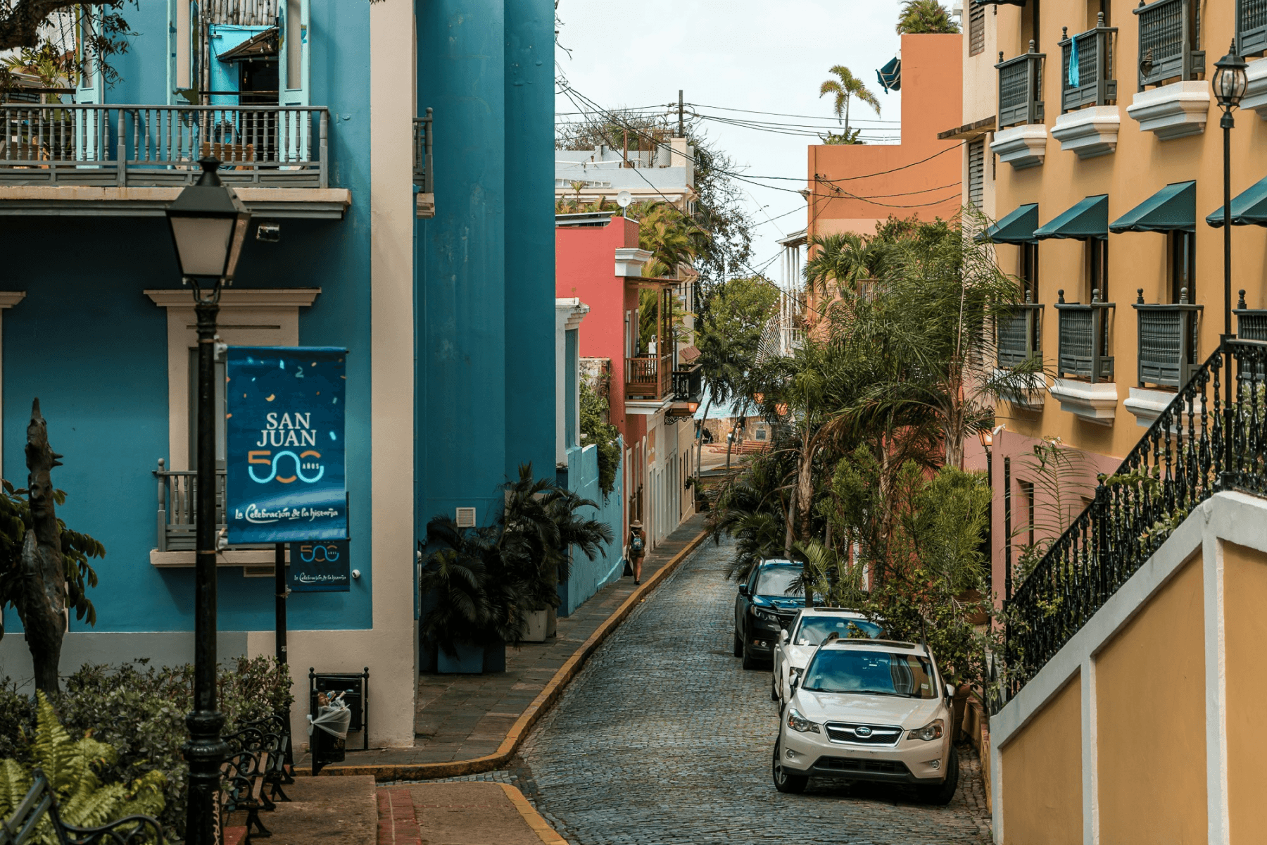 A colorful cobblestone street in Old San Juan, Puerto Rico, lined with vibrant colonial buildings in blue, pink, and yellow, with a San Juan 500th anniversary banner visible.