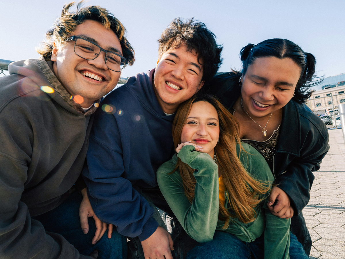 Four young friends huddled close together, laughing and smiling at the camera outdoors on a sunny day, with lens flare and a clear sky in the background.
