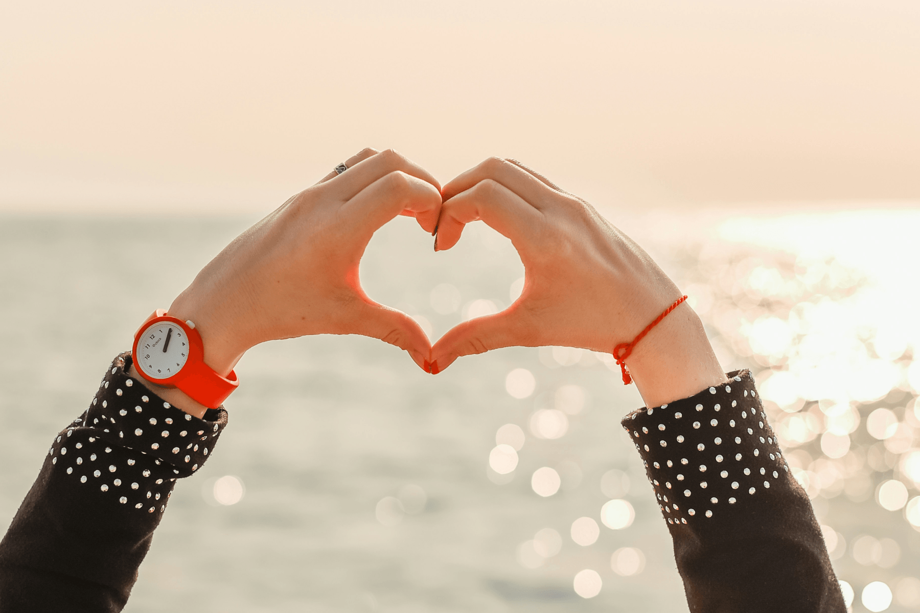 A person making a heart shape with their hands against a sun-reflecting ocean, wearing a red watch and polka-dot sleeves.