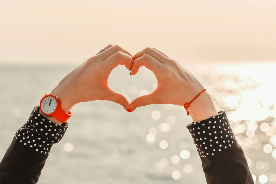 A person making a heart shape with their hands against a sun-reflecting ocean, wearing a red watch and polka-dot sleeves.