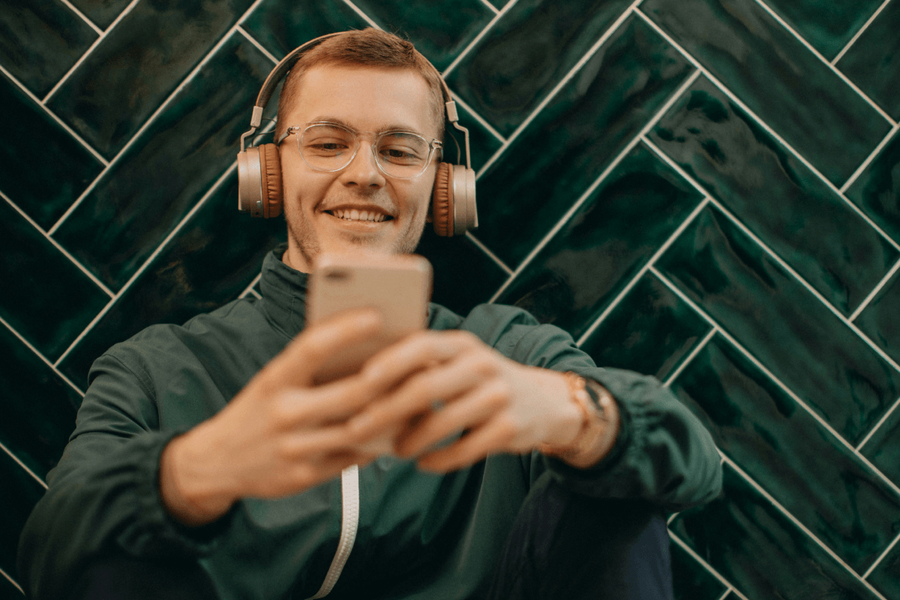 A young man with glasses and headphones smiling while looking at his phone against a green herringbone tile wall.