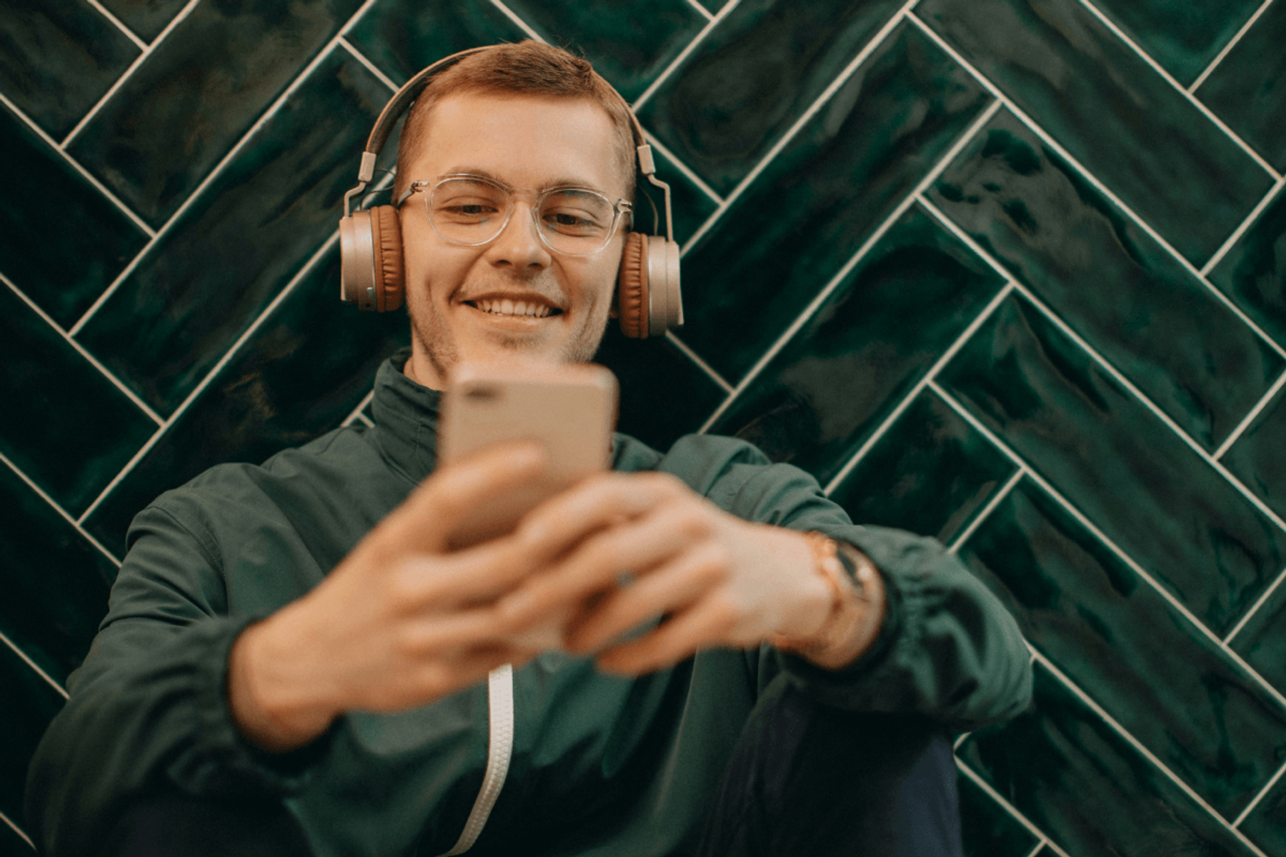 A young man with glasses and headphones smiling while looking at his phone against a green herringbone tile wall.
