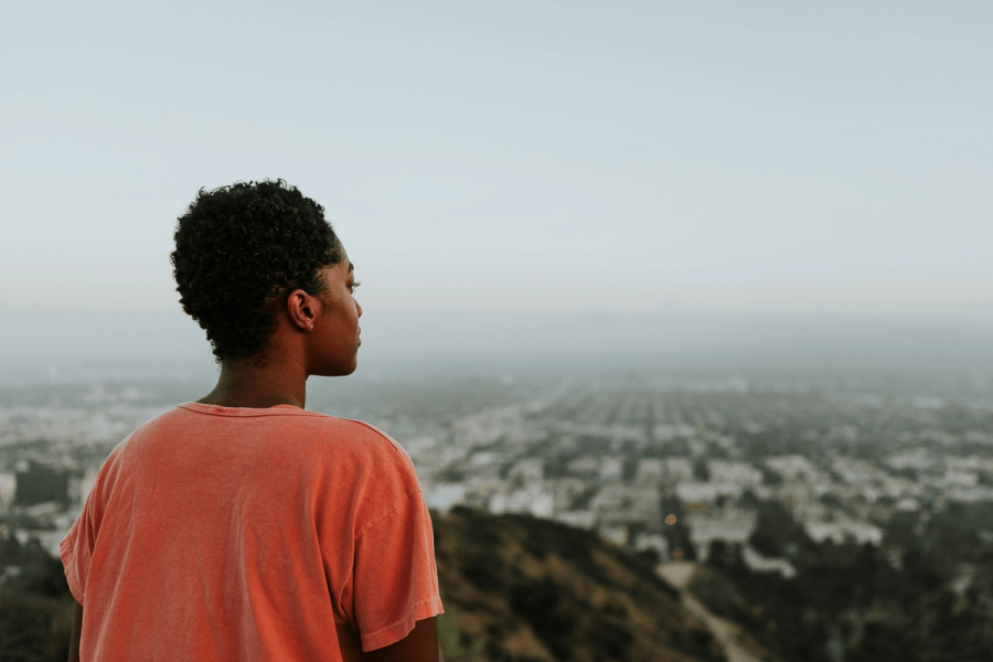 A person seen from behind in a pink t-shirt, gazing out over a sprawling city from a hilltop on a hazy day.