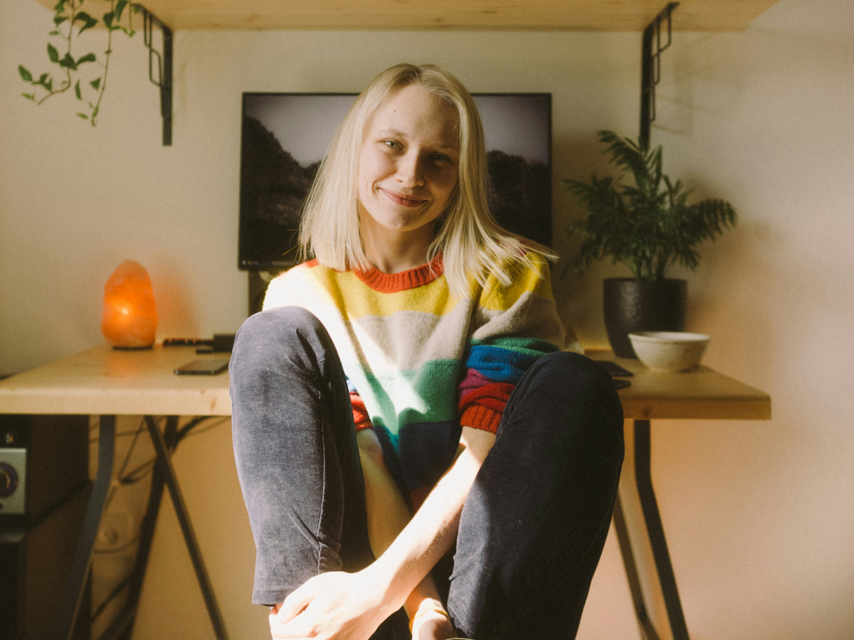 A young woman in a rainbow striped sweater sitting relaxed on the floor in front of a wooden desk, smiling warmly in soft natural light with houseplants and a salt lamp in the background.