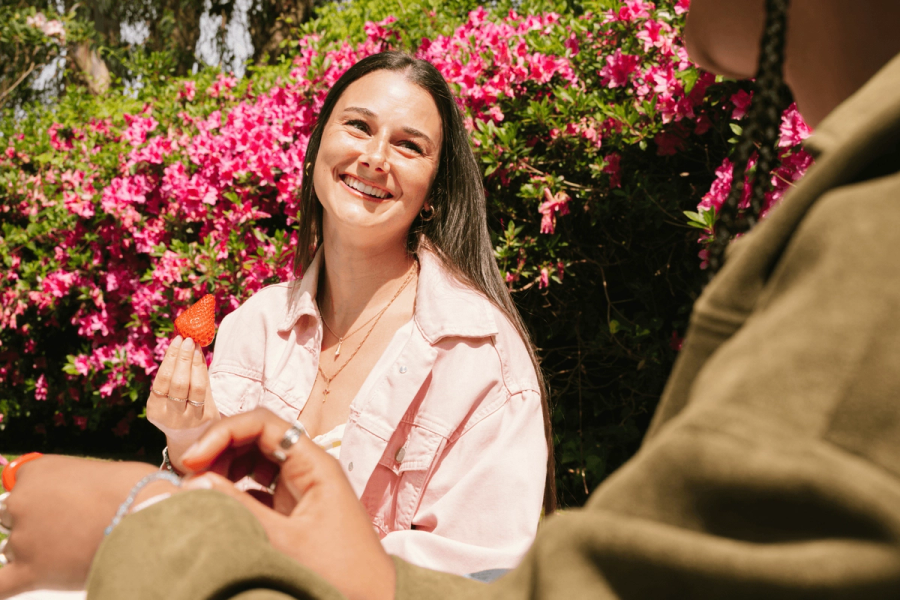 Claude responded: A smiling young woman holding a strawberry outdoors, sitting in front of vibrant pink flowering bushes while talking with someone.A smiling young woman holding a strawberry outdoors, sitting in front of vibrant pink flowering bushes while talking with someone.