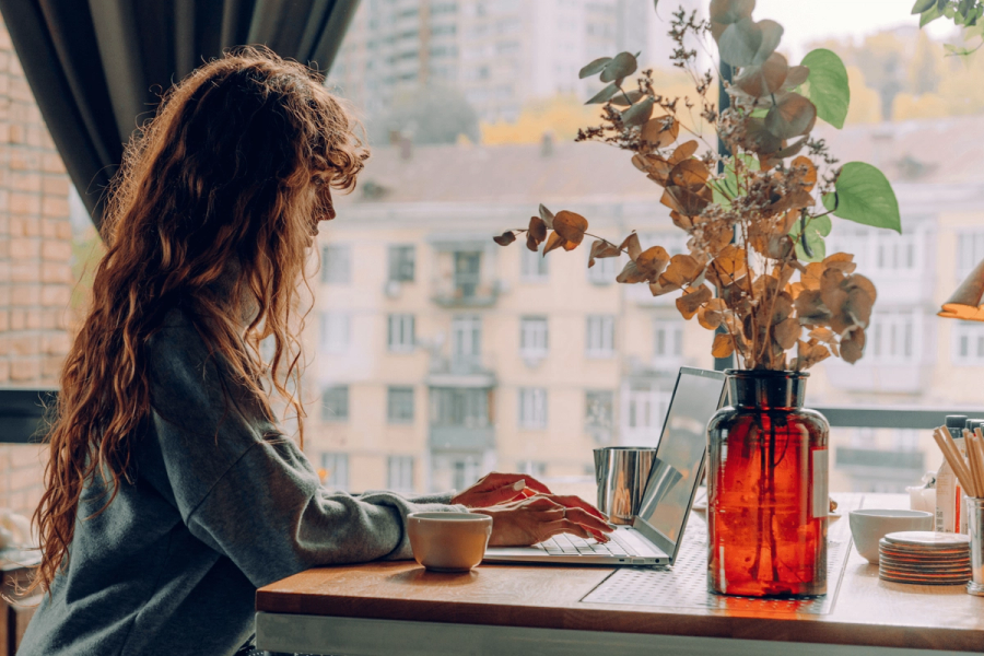 A woman with long curly red hair typing on a laptop at a café window, with a dried eucalyptus arrangement and a cup of coffee beside her.