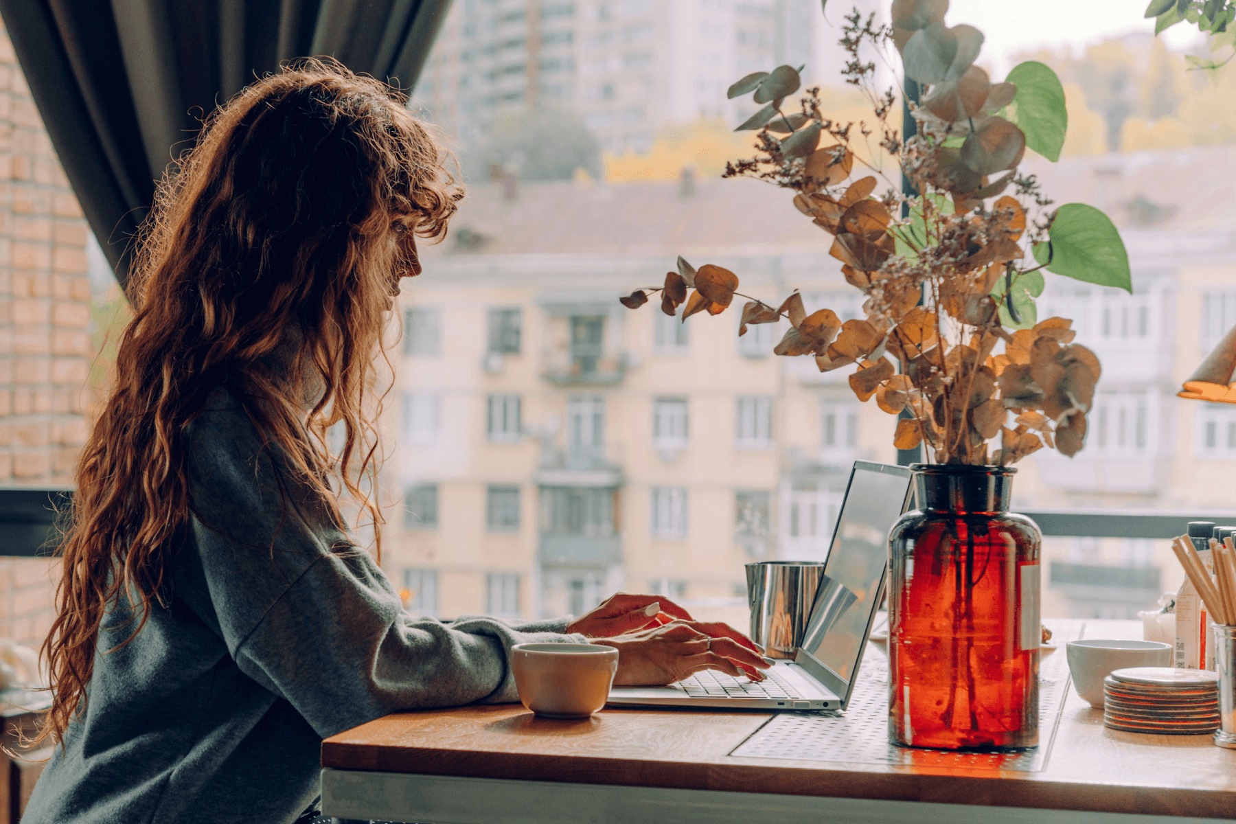 A woman with long curly red hair typing on a laptop at a café window, with a dried eucalyptus arrangement and a cup of coffee beside her.