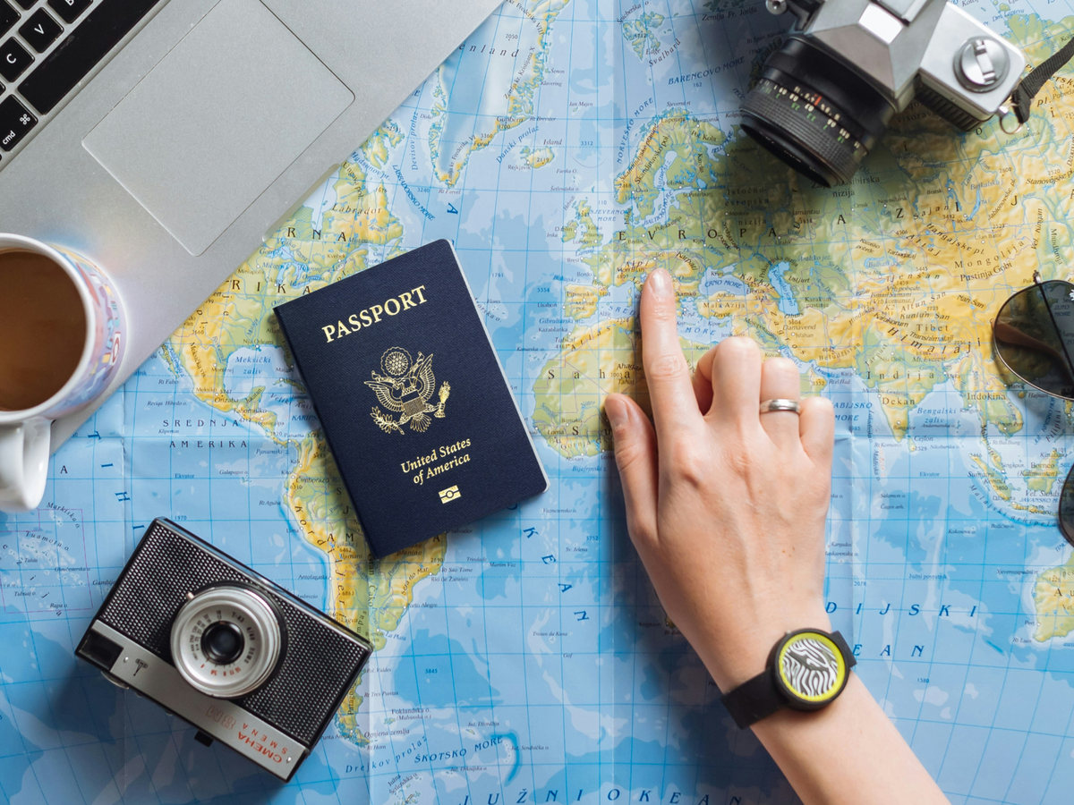 Overhead flat-lay of a world map with a US passport, two cameras, sunglasses, and a coffee cup arranged around it, while a hand wearing a watch points to a location in Europe.