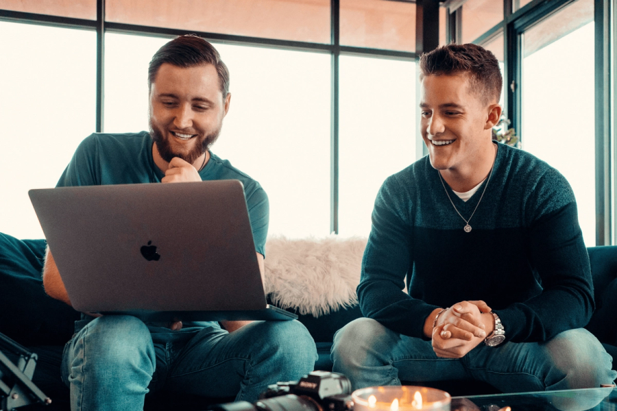 Two men smiling while looking at a MacBook together, seated on a couch in a bright, modern space.