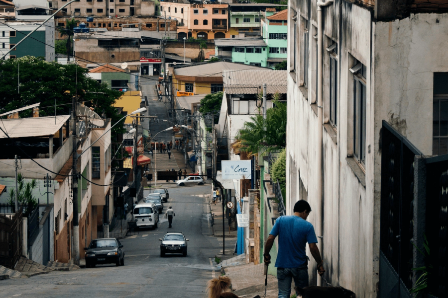 A hilly Brazilian street scene with colorful buildings, cars, and a man walking a dog in the foreground.