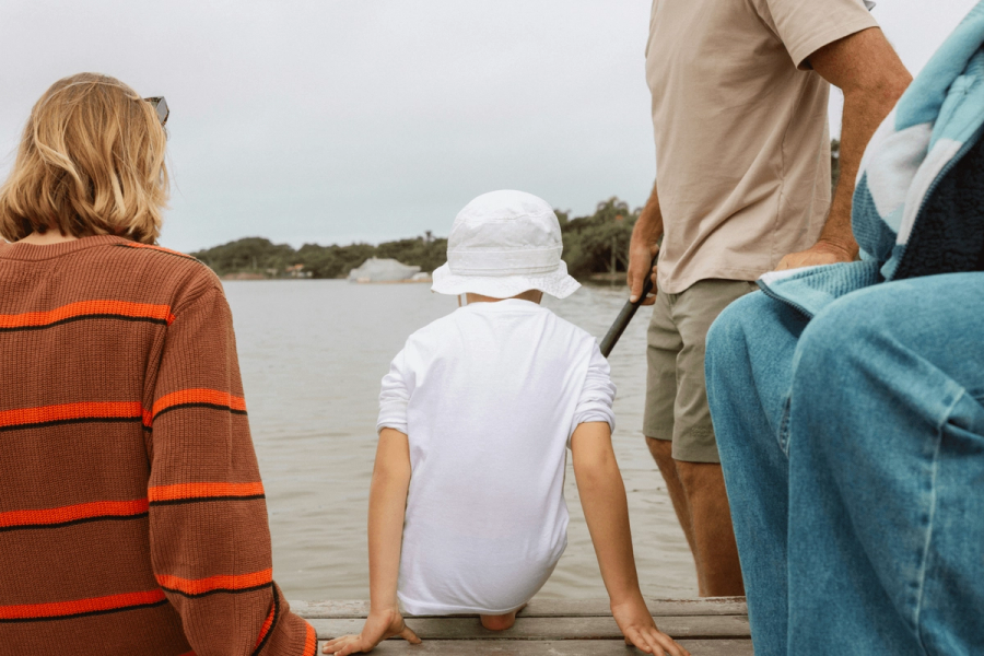 A family seen from behind sitting and standing on a dock overlooking a calm river, with a child in a white sun hat seated in the middle.A family seen from behind sitting and standing on a dock overlooking a calm river, with a child in a white sun hat seated in the middle.