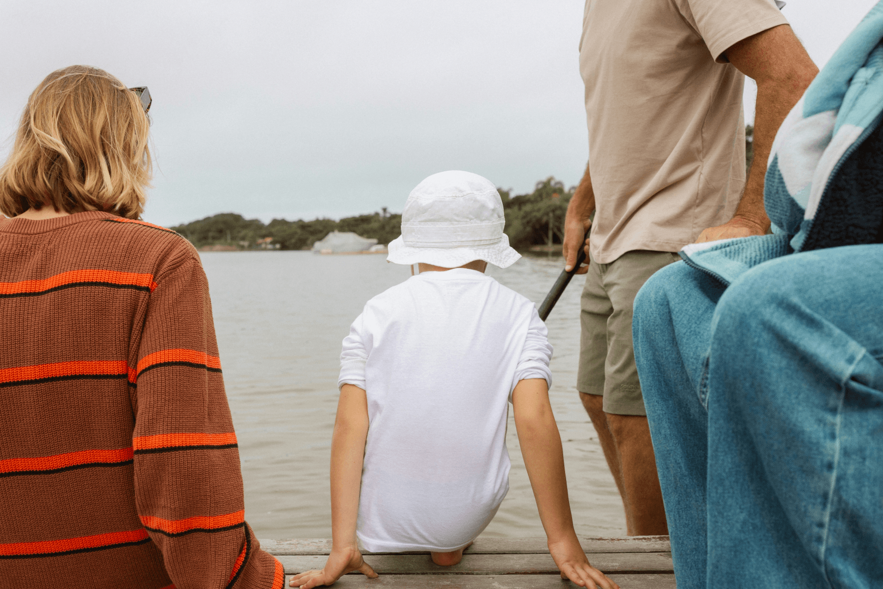 A family seen from behind sitting and standing on a dock overlooking a calm river, with a child in a white sun hat seated in the middle.A family seen from behind sitting and standing on a dock overlooking a calm river, with a child in a white sun hat seated in the middle.