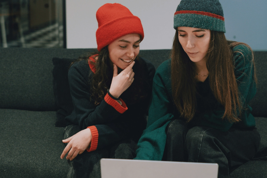 Two women wearing beanies sitting together on a couch, smiling and looking at a laptop.