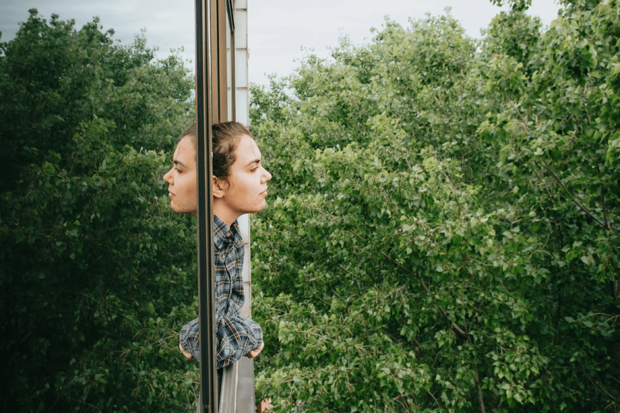 A person in a flannel shirt looking pensively to the side, with their reflection visible in a glass window, surrounded by lush green trees.