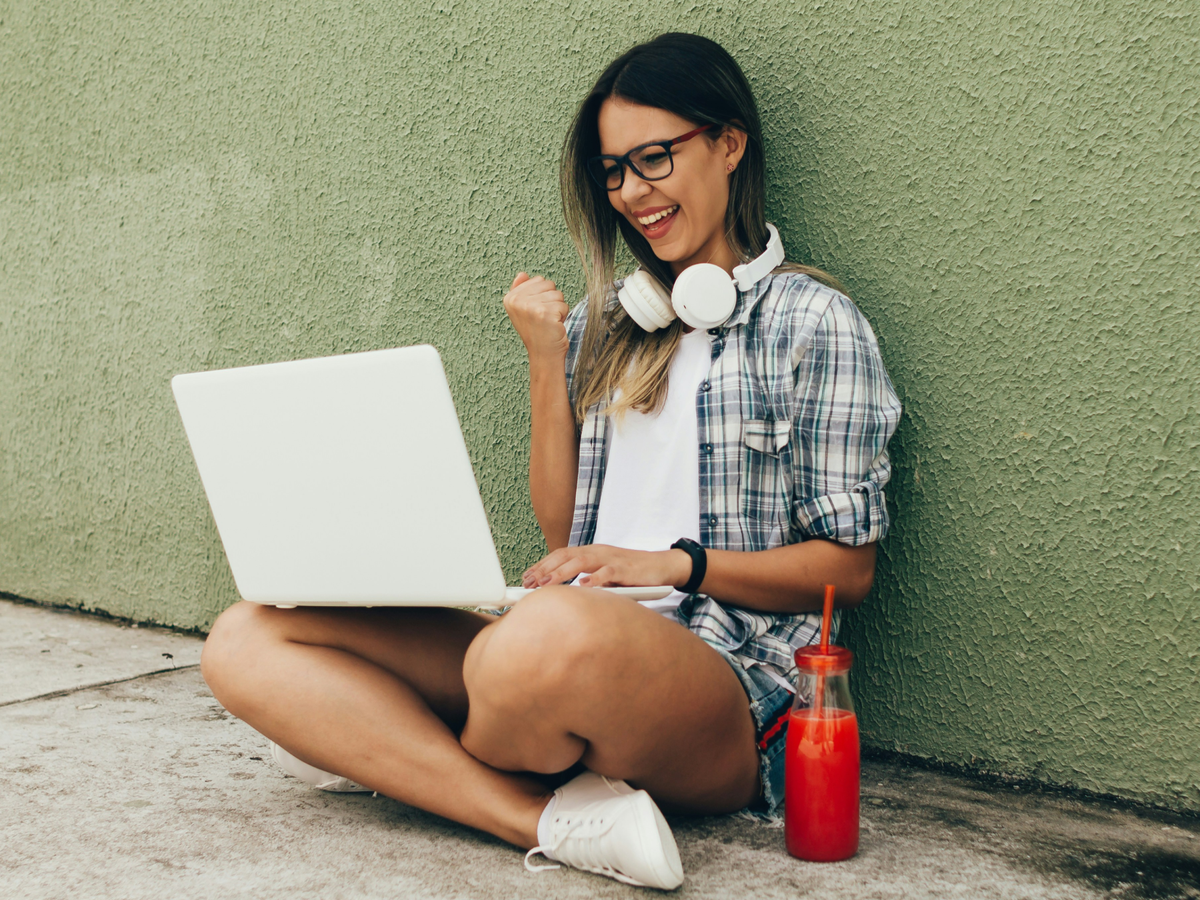 A young woman sitting cross-legged on the ground against a green wall, smiling excitedly at her laptop with headphones around her neck and a red drink beside her.