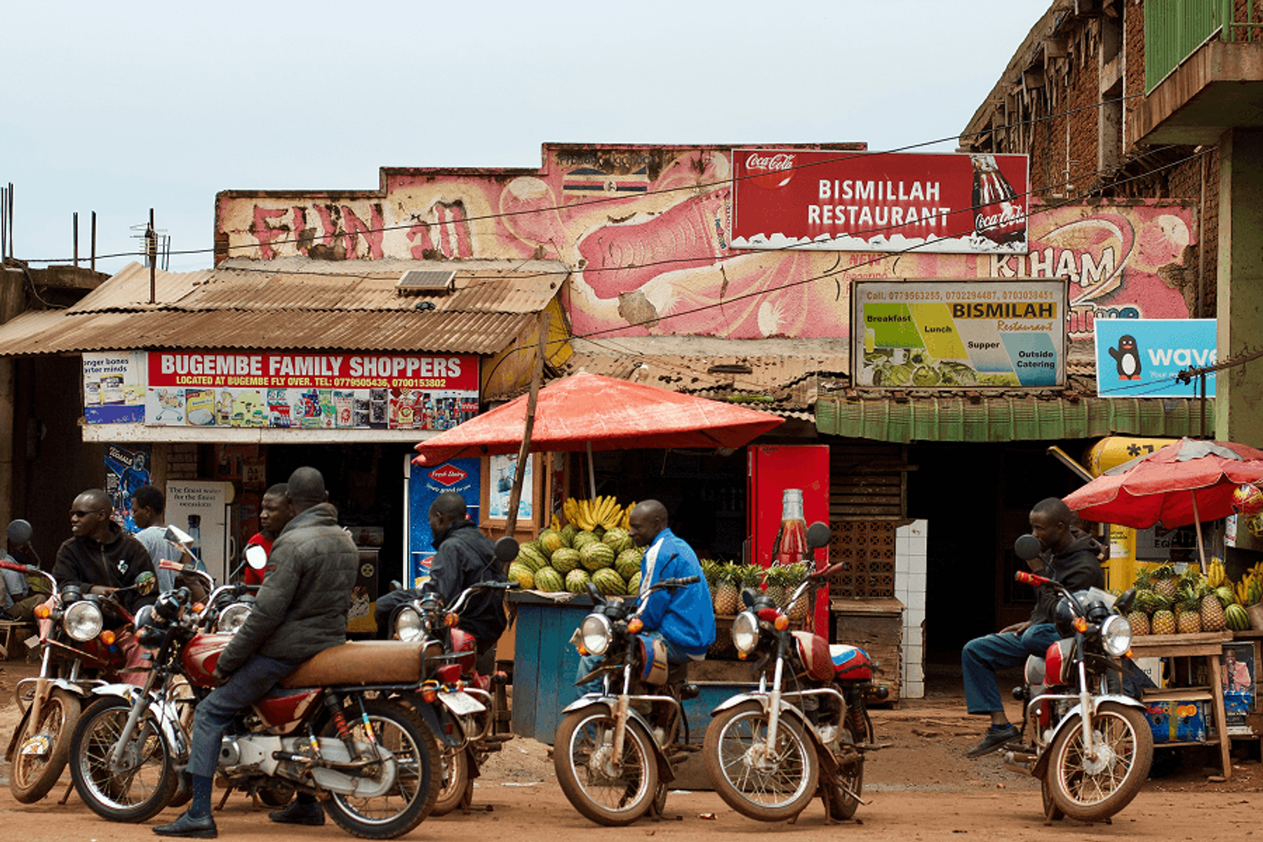 A busy Ugandan street scene with motorcycle taxis, fruit vendors, and colorful storefront signs including a restaurant and family shop.