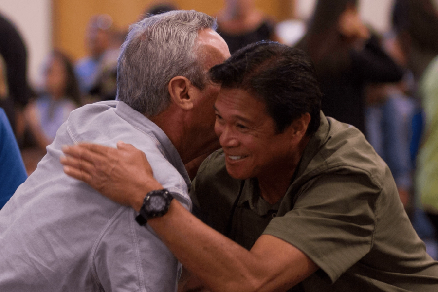 Two men embracing warmly in what appears to be a church gathering, with a crowd of people visible in the background.Two men embracing warmly in a church gathering, with a crowd of people visible in the background.