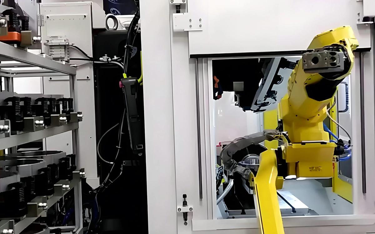 Yellow industrial robotic arm reaching toward the open enclosure of a CNC machine, with a rack of fixtures or tools on the left.