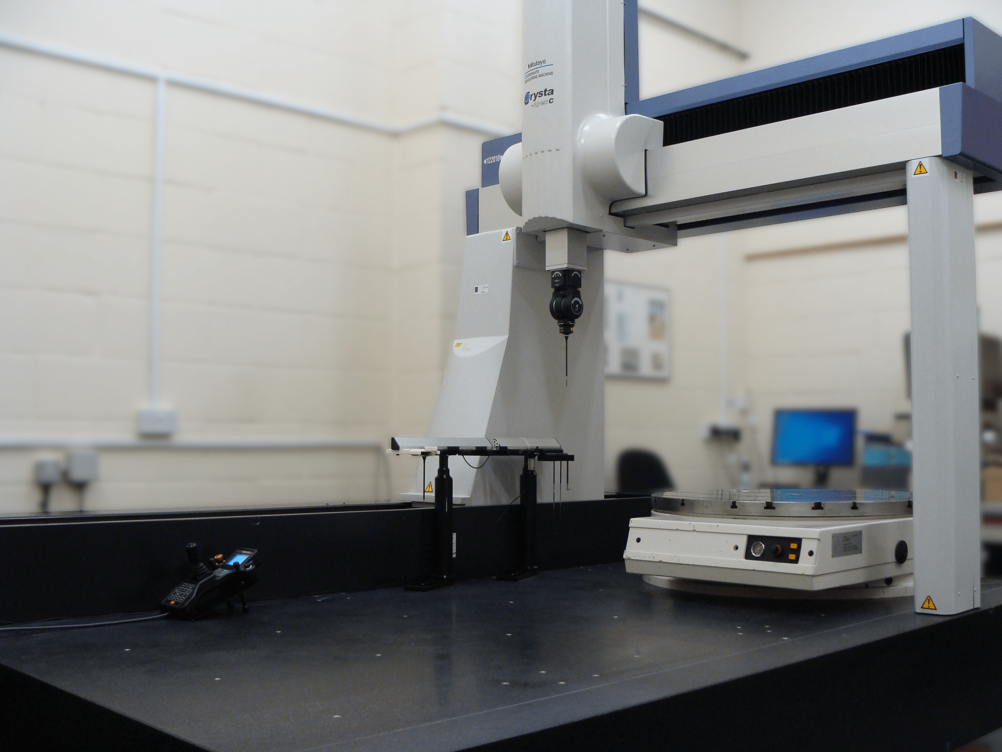 Five-axis scanning coordinate measuring machine with a vertical measurement head and rotary table in an inspection room.